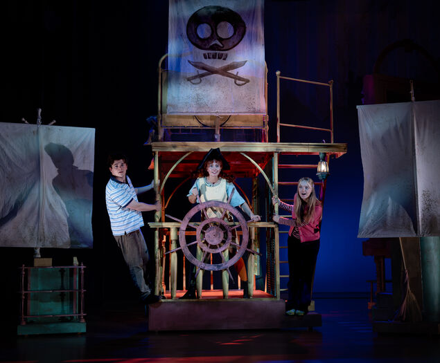 Drie kinderen spelen op een theaterdecor met een schip, waar één kind aan het roer staat en de anderen poseren.