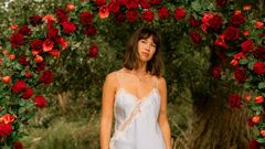 MEAU staat onder een boog van rode rozen in een bosachtige omgeving, gekleed in een licht topje en witte broek.