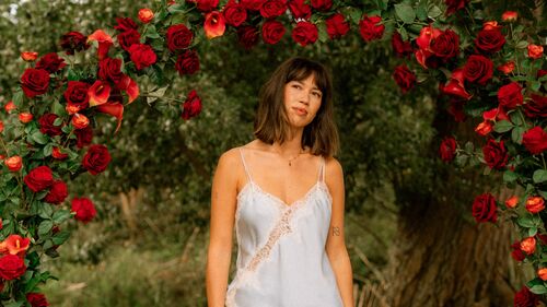 MEAU staat onder een boog van rode rozen in een bosachtige omgeving, gekleed in een licht topje en witte broek.