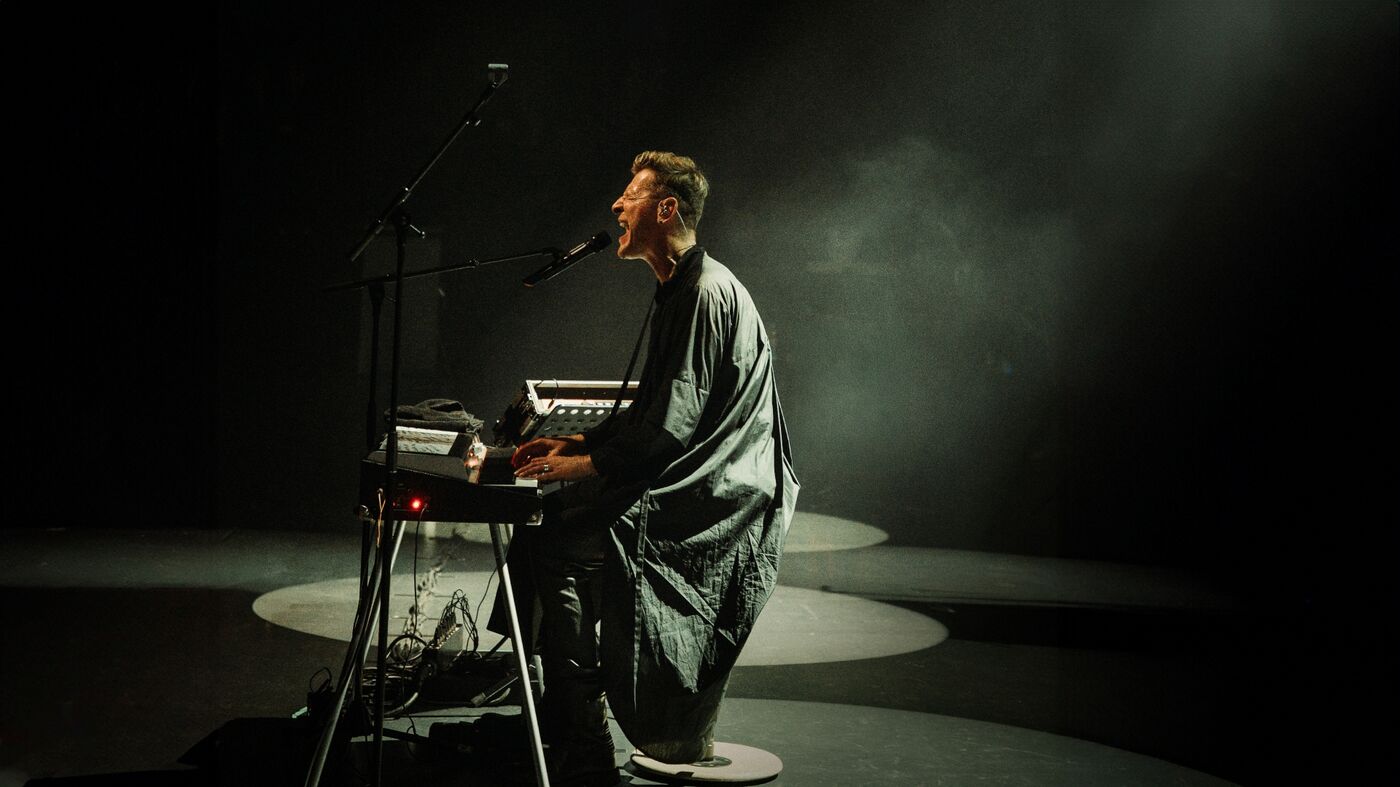 Performer (Ozark Henry) zit bij een keyboard en microfoon in een donkere ruimte, belicht door een smalle lichtbundel.