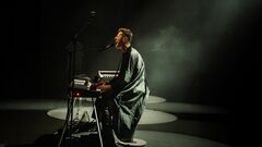 Performer (Ozark Henry) zit bij een keyboard en microfoon in een donkere ruimte, belicht door een smalle lichtbundel.