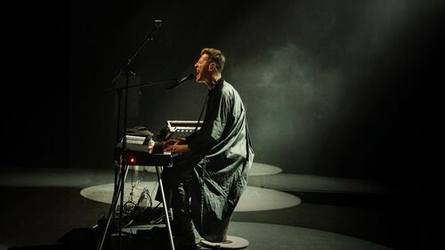 Performer (Ozark Henry) zit bij een keyboard en microfoon in een donkere ruimte, belicht door een smalle lichtbundel.