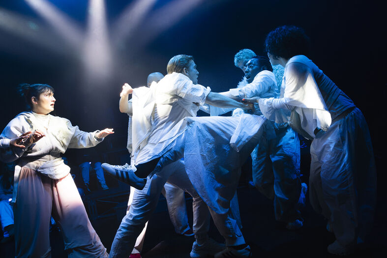 Groep dansers in witte kostuums voert een dynamische, samenwerkende choreografie uit op een podium met spotlichten.