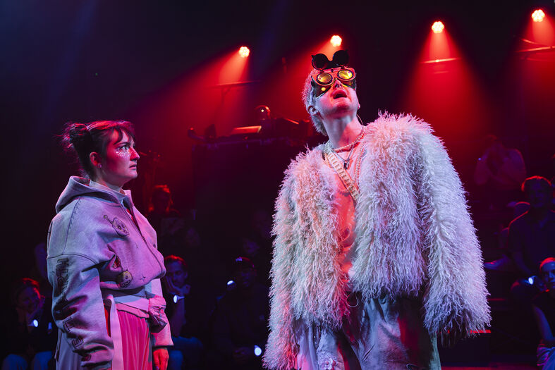 Twee acteurs in theatrale kostuums staan in een kleurrijke verlichte scène met een publiek op de achtergrond.