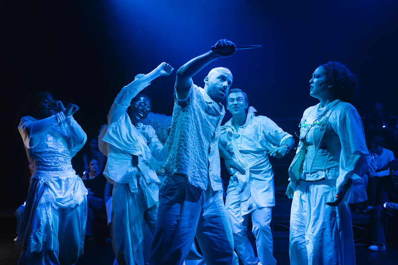 Groep dansers in kostuums voert een energieke choreografie uit op een podium met blauwe verlichting.