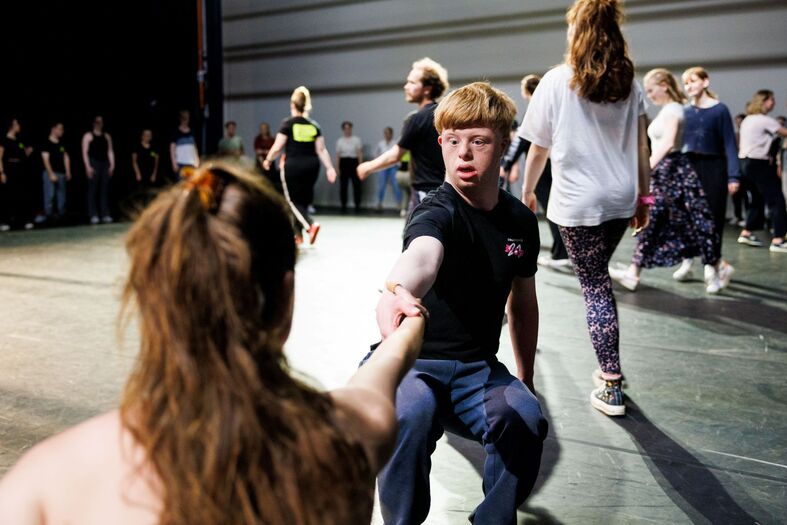 Groep dansers in een repetitie, met een jongen die een hand uitsteekt naar een meisje terwijl anderen achter hen aan het dansen zijn.