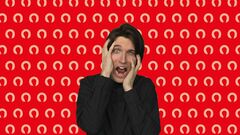 Persoon met een verontruste uitdrukking en handen op het hoofd, voor een rode achtergrond met witte, herhalende logo's.