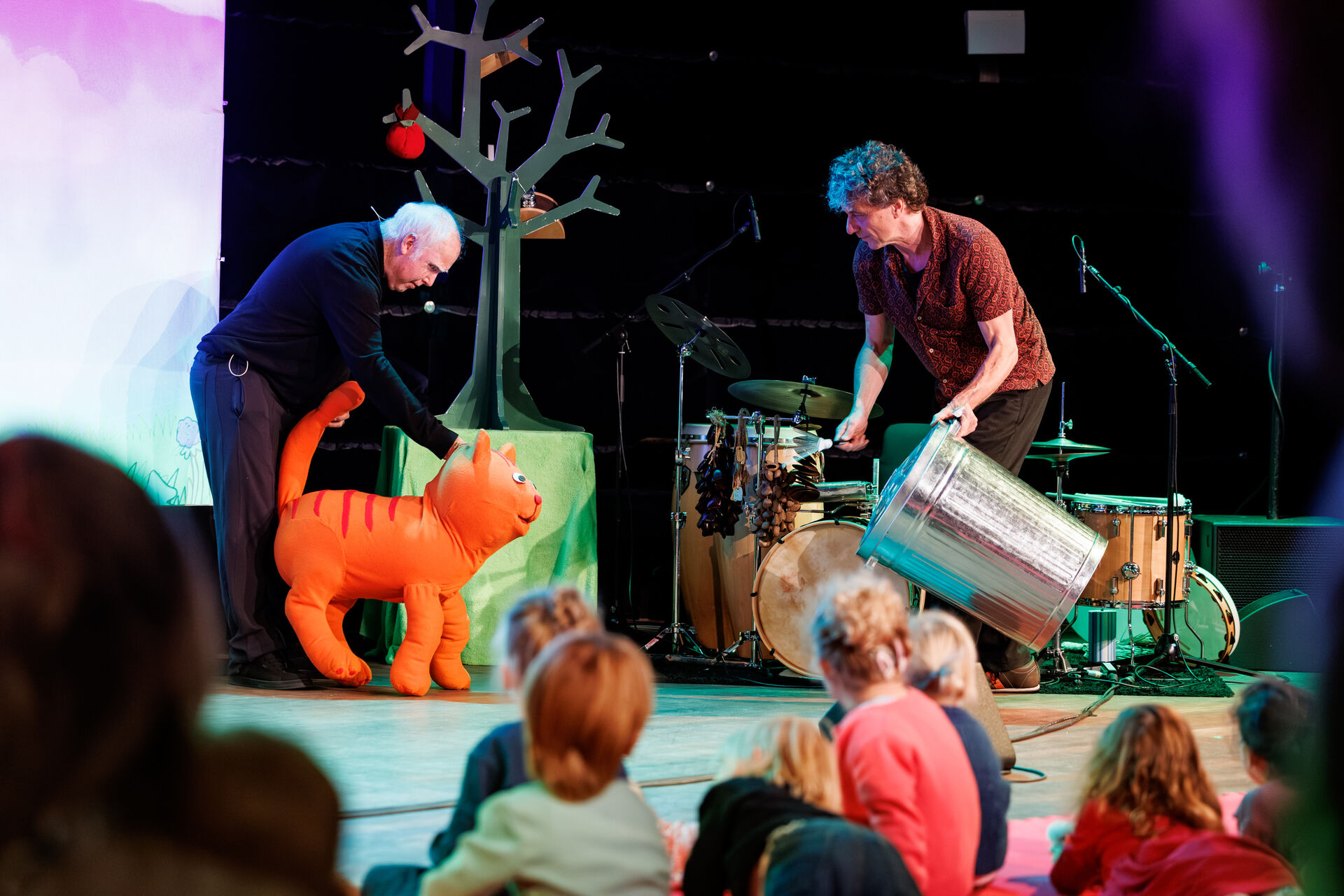 Twee performers op een podium voeren een voorstelling op voor een groep kinderen, waaronder een grote, oranje kat.