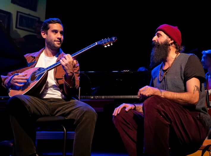 Twee muzikanten zitten op een podium, de ene speelt een gitaar, de andere luistert aandachtig.