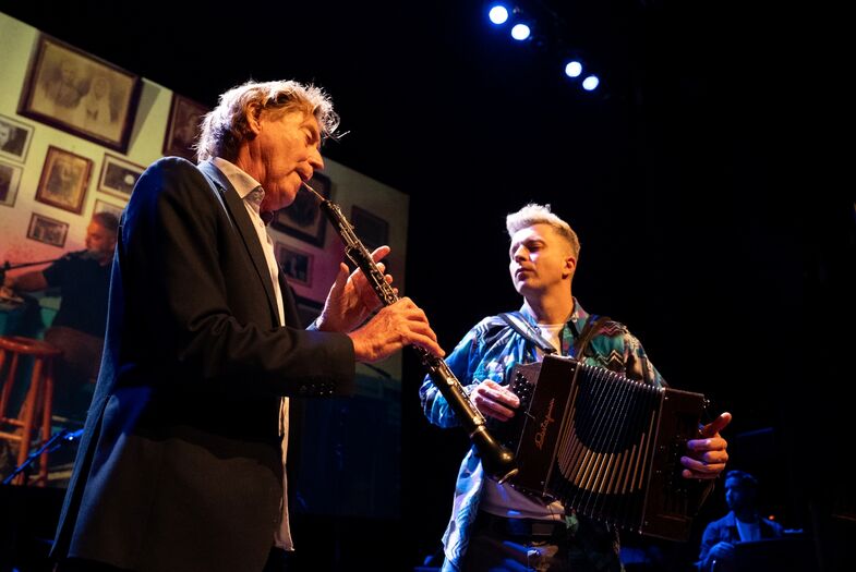 Twee muzikanten voeren een live optreden uit, één op de klarinet en de ander op de accordeon, met een podiumdecor.