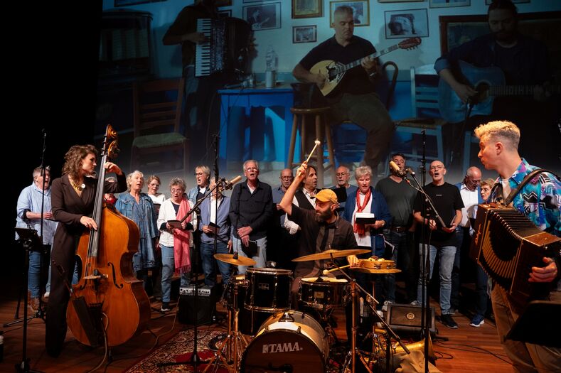Een muzikale uitvoering met een koor en bandleden, inclusief een drummer en een gitarist, op een verlichte podium.