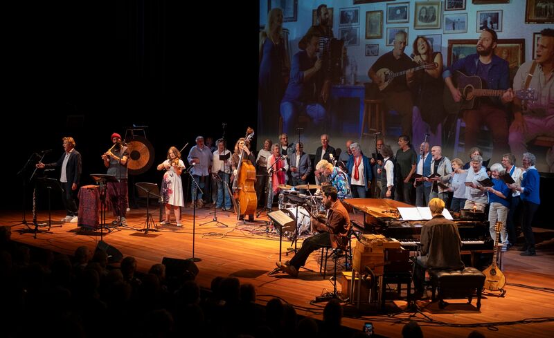 Groep muzikanten en zangers op het podium, met een projector op de achtergrond die foto's weergeeft.