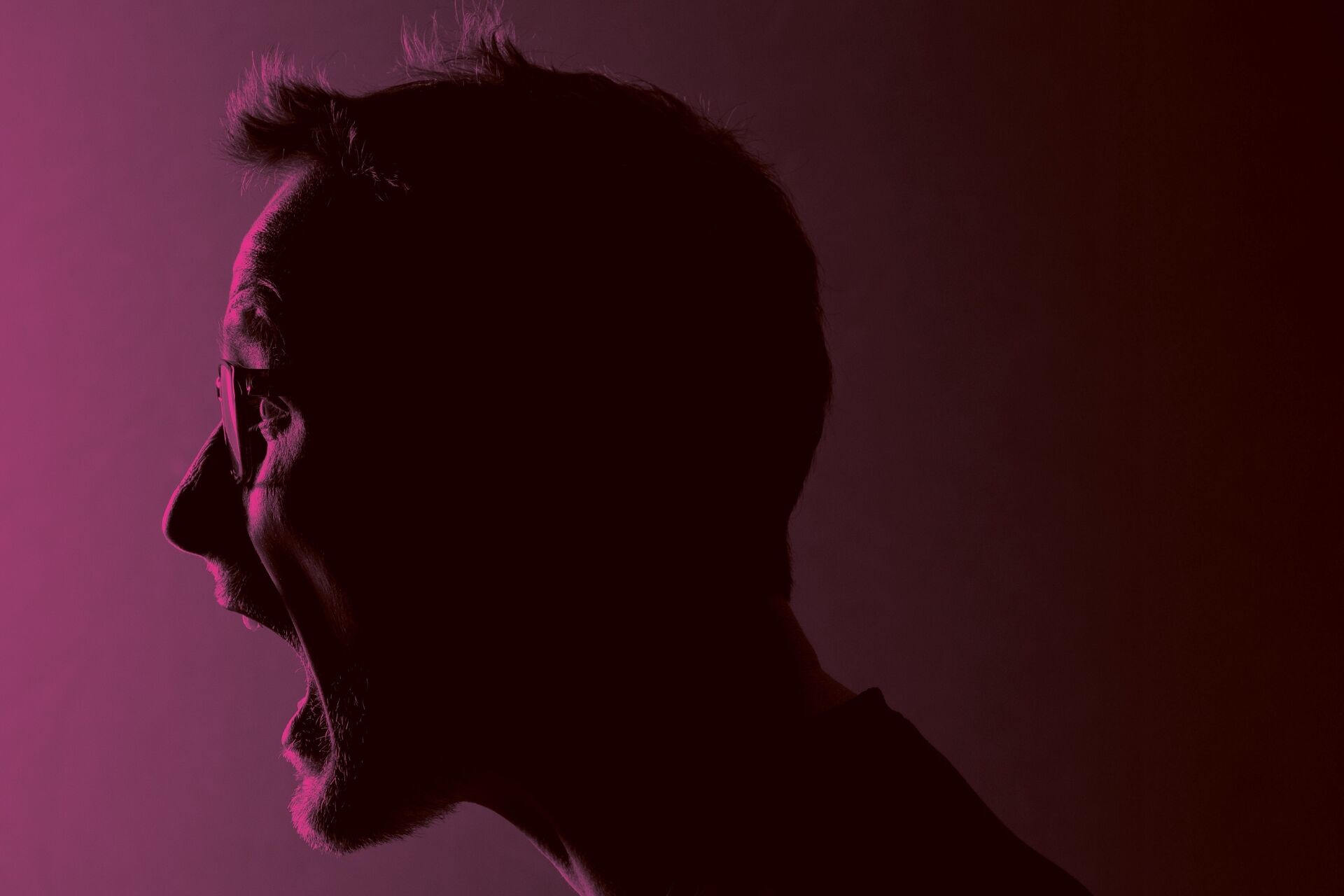Zijprofiel van een persoon die met wijd open mond schreeuwt, belicht in roze en donkerrood tegen een donkere achtergrond.