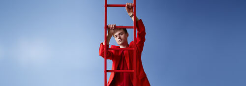 Luuk Ransijn in felrode kleding klimt aan een rode ladder tegen een heldere blauwe lucht.