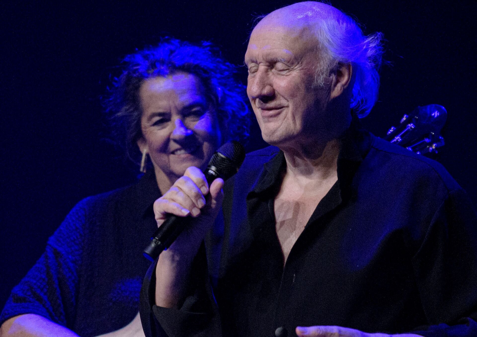 Twee personen op een donker podium, één met microfoon in de hand.