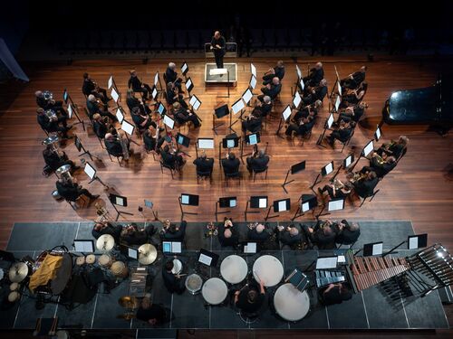 Bovenaanzicht van een orkest dat repeteert, met diverse instrumenten en muzikanten in een concertzaal.