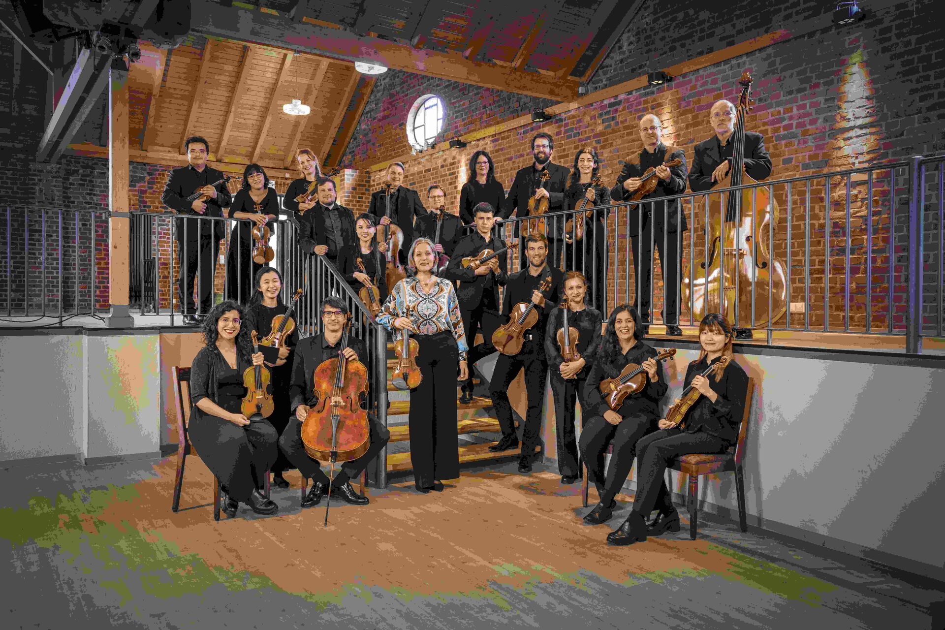 Een orkest bestaande uit 18 muzikanten, poserend op een trap met verschillende instrumenten in een sfeervolle ruimte.