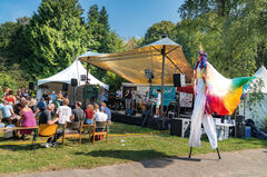 Een buitenfestival met een podium, muzikanten en een publiek, omringd door kleurrijke kraampjes en decoraties.