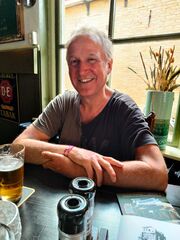 Een man met grijs haar zit aan een tafeltje in een café, lachend met een biertje voor zich.