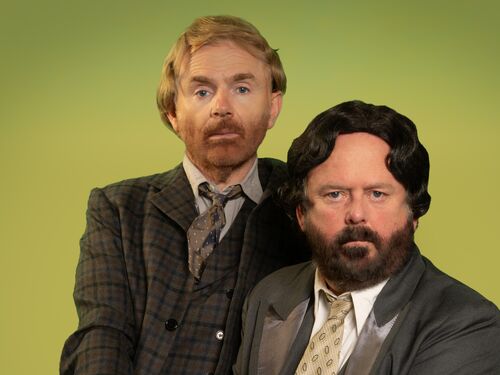 Twee mannen in vintage kostuums zitten op een groene achtergrond, met verschillende kapsels en gezichtsbeharing.