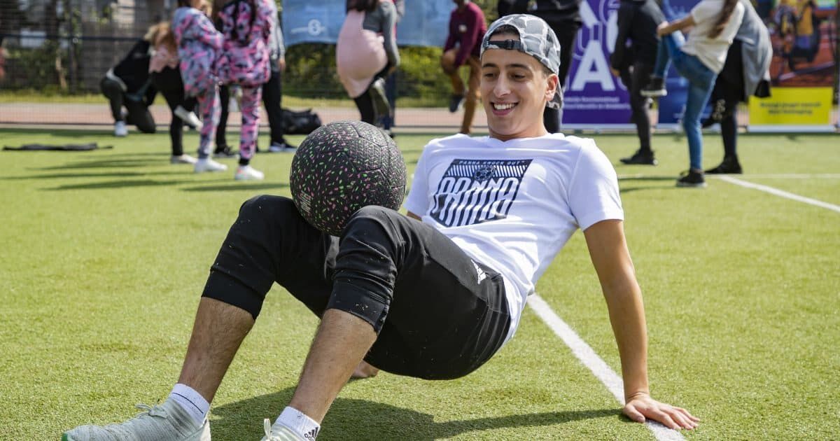 Jongeman zittend op grasveld, genietend van een sportactiviteit met een bal tussen zijn benen. Achtergrond met mensen en een evenement.