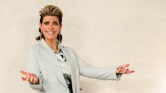 Vrouw in een lichtgrijze blazer staat met uitgestrekte armen en een glimlach voor een neutrale achtergrond.