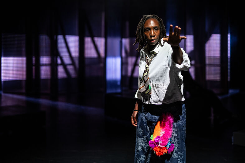 Persoon in theatrale outfit met kleurrijke accessoires staat in een donkere ruimte en steekt hand naar voren.