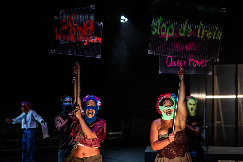 Drie performers met kleurrijke maskers houden protestborden omhoog in een theatrale setting.