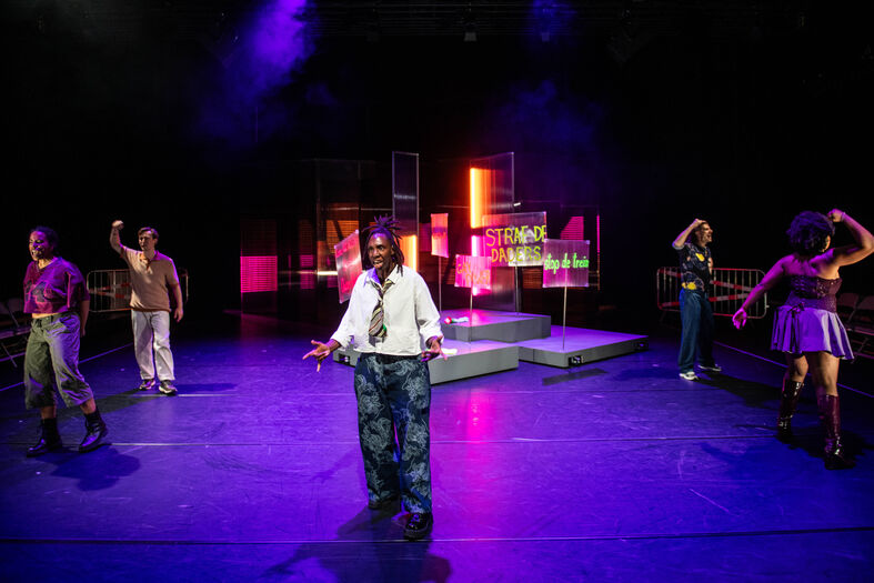 Een theaterstuk met vijf performers op een verlichte scène in kleurrijke outfits.