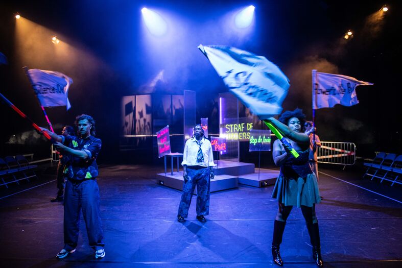 Drie acteurs in een theater zetten kleurrijke vlaggen in de lucht op een verlicht podium.