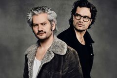 Twee personen staan rug aan rug in donkere jassen tegen een neutrale grijze achtergrond.
