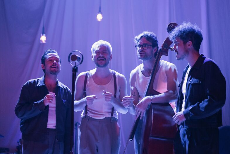 Vier muzikanten staan op een podium, met een microfoon en een contrabas, onder een paar sfeervolle lampen.