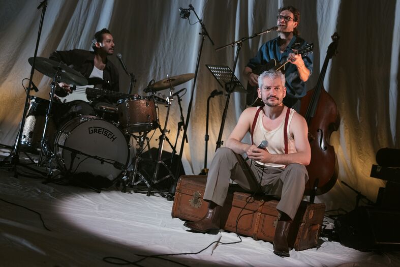 Muzikanten op een podium: een man speelt gitaar, een ander zit op kisten en de drummer speelt op een drumstel.