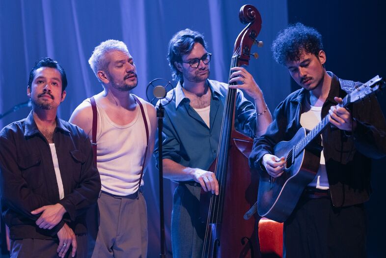 Vier muzikanten staan op een podium, twee zingen, terwijl één een contrabas en één een gitaar bespeelt.
