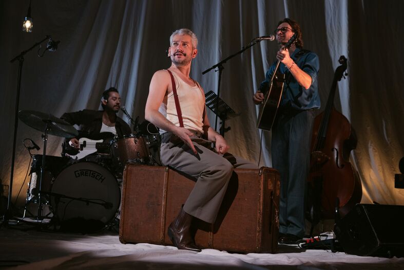 Drie muzikanten voeren een live optreden uit, met een zanger op een koffer en bandleden op drums en contrabas.