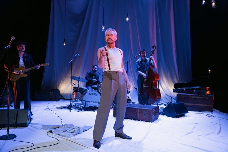 Man in casual kleding staat op een podium met een band, concertsetting met lichten en een witte achtergrond.