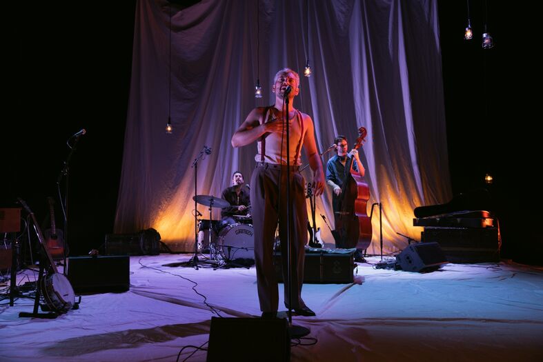Zanger op het podium met muzikanten, een bassist en een drummer, tegen een sfeervolle achtergrond van verlichting.