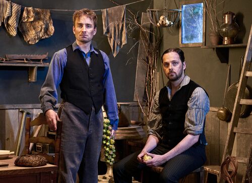 Twee mannen (Rundfunk) in historische kleding staan in een sfeervolle, verouderde kamer met een houten ladder en een kledinglijn.