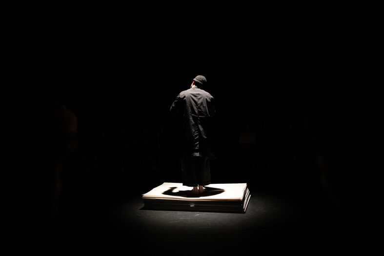 Een geklede performer staat op een verhoogd platform in een smalle lichtcirkel, omringd door een duister publiek.