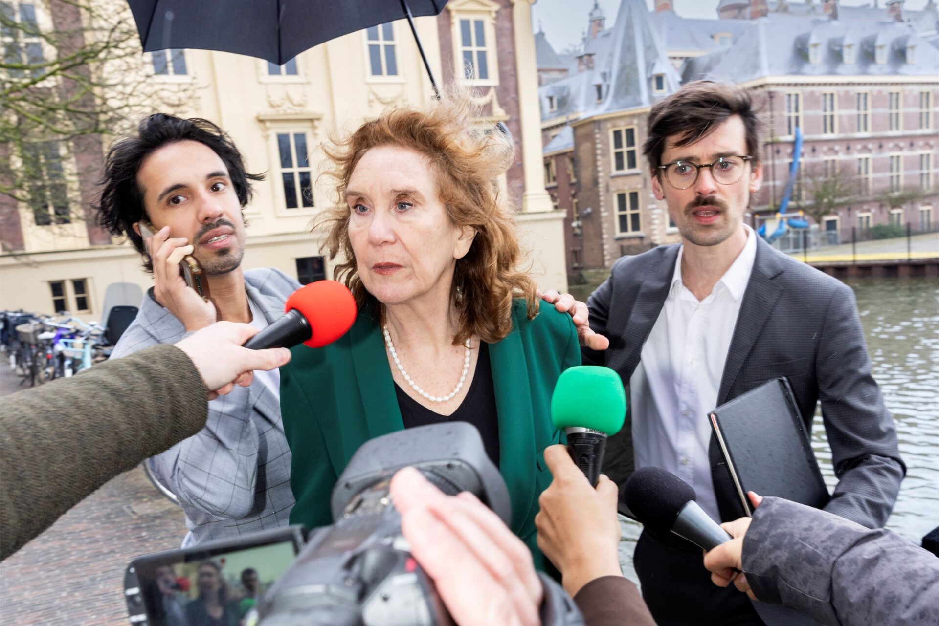 Drie personen lopen buiten langs het Binnenhof terwijl meerdere microfoons naar hen worden uitgestoken.