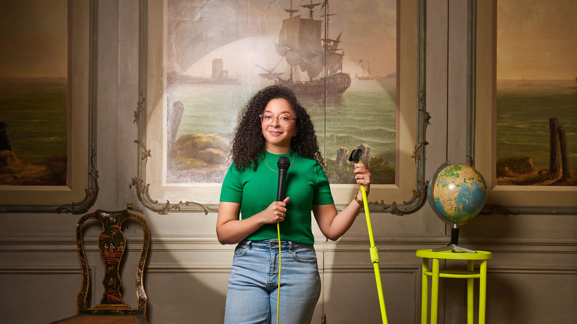 Persoon (Wina Ricardo) staat in een klassieke kamer met schildering van een zeilschip, houdt een microfoon en een felgele microfoonstandaard vast, naast een antieke stoel en een kleine tafel met wereldbol.