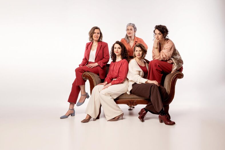 Vijf vrouwen in stijlvolle kleding poseren samen op een bank voor een neutrale achtergrond.