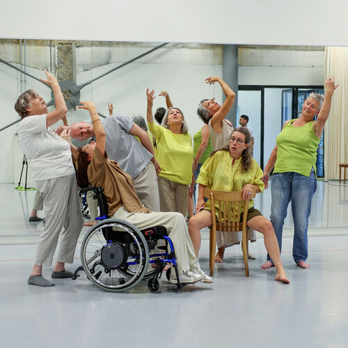 Een groep dansers, waaronder een persoon in een rolstoel, in een studio, die gezamenlijk een expressieve houding aannemen.