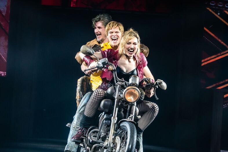 Drie acrobaten op een motor, poseren in een theatrale omgeving met een energieke uitstraling.