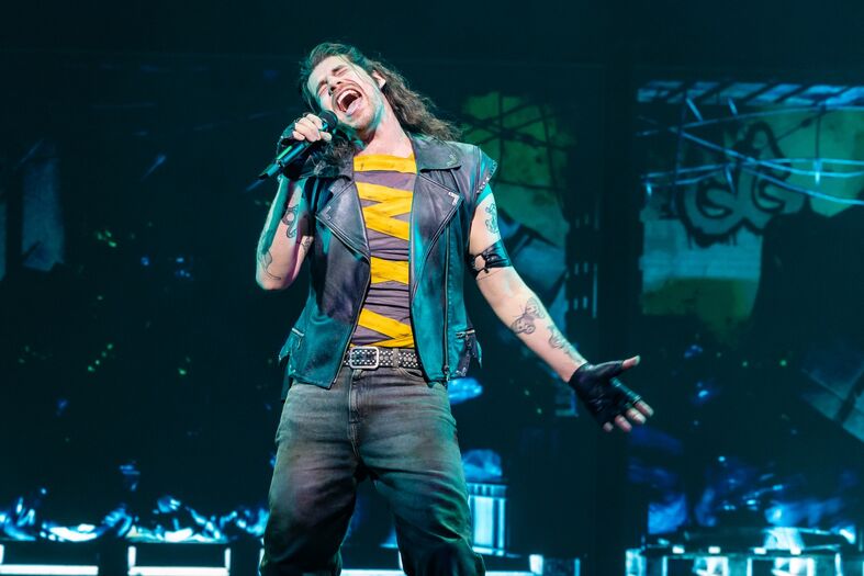 Zanger met een leren vest en lange haren die oplicht in een podiumlicht tijdens een liveoptreden.