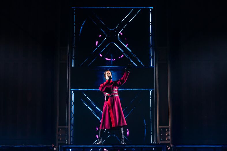 Een performer in een lange, rode outfit staat op een podium met een theatrale achtergrond en lichteffecten.