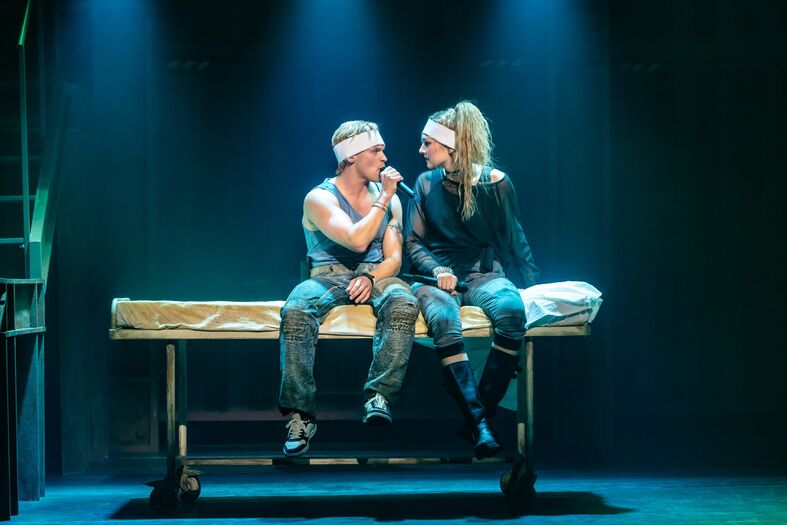 Twee acteurs zitten naast elkaar op een bed in een theater, verlicht door blauwe spots.