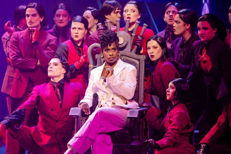 Een groep artiesten in rode kostuums rondom een man in witte kleding op een troon, op een kleurrijk podium.