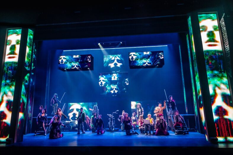 Theater voorstelling met artiesten op het podium, omgeven door visuele projecties en kleurrijke verlichting.