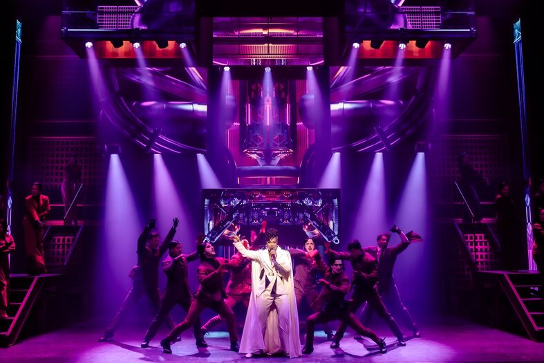 Een muzikale voorstelling met een artiest in het midden, omringd door dansers onder kleurrijke verlichting.